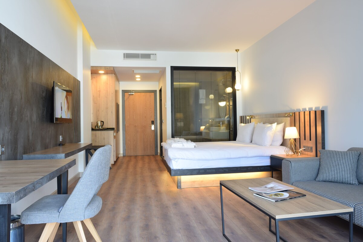Een moderne hotelkamer met een houten vloer, een bed met witte lakens, een grijze fauteuil, een houten salontafel en een klein tafeltje met een lamp.