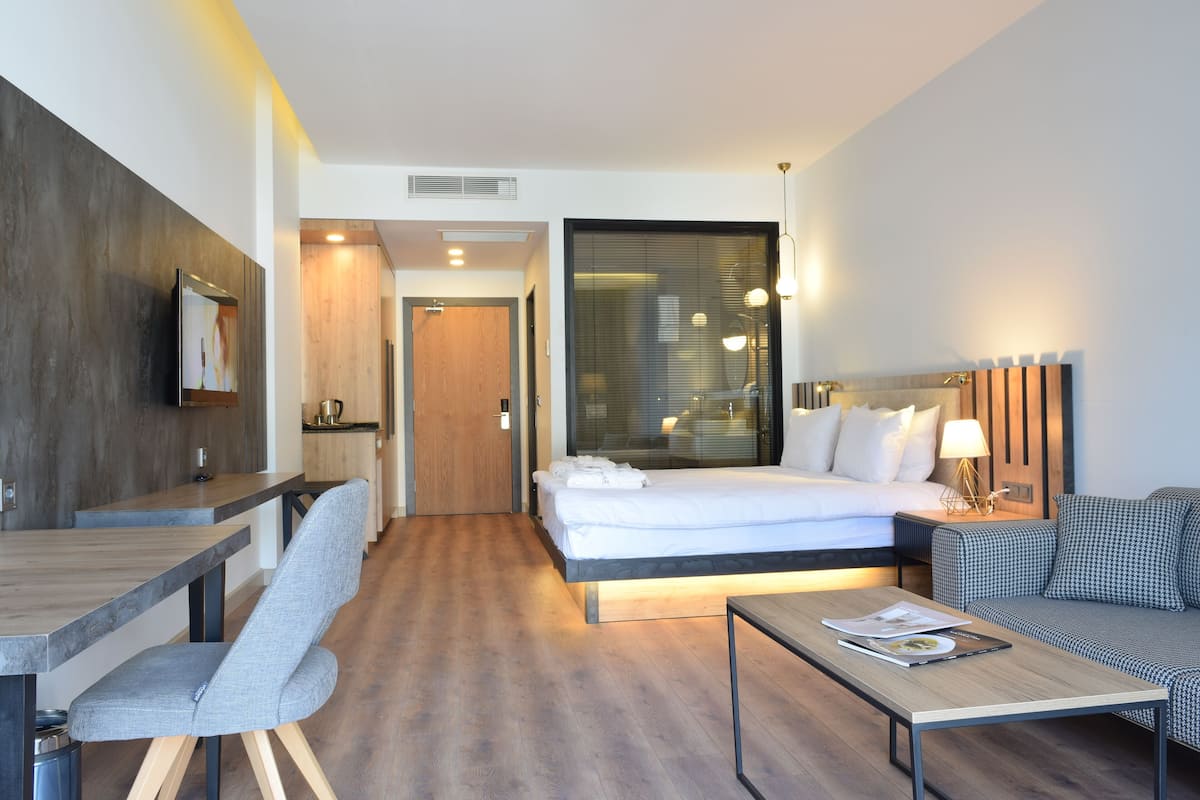 Une chambre d’hôtel moderne avec un plancher en bois, un lit recouvert de draps blancs, un fauteuil rembourré de couleur grise, une table basse en bois et une petite table avec une lampe.