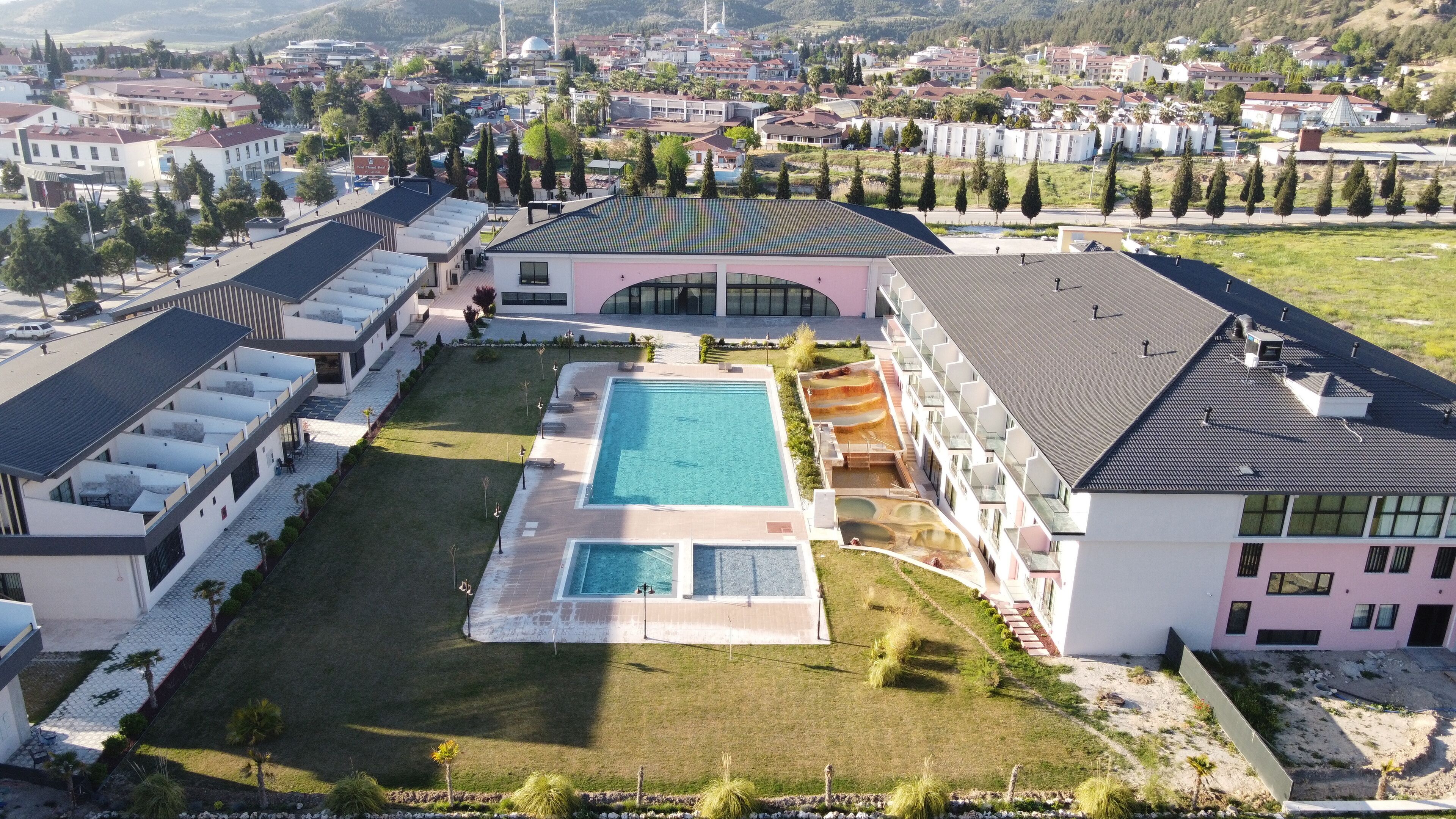 An aerial view of a modern hotel with a central swimming pool, multiple balconies, and a surrounding landscaped area.