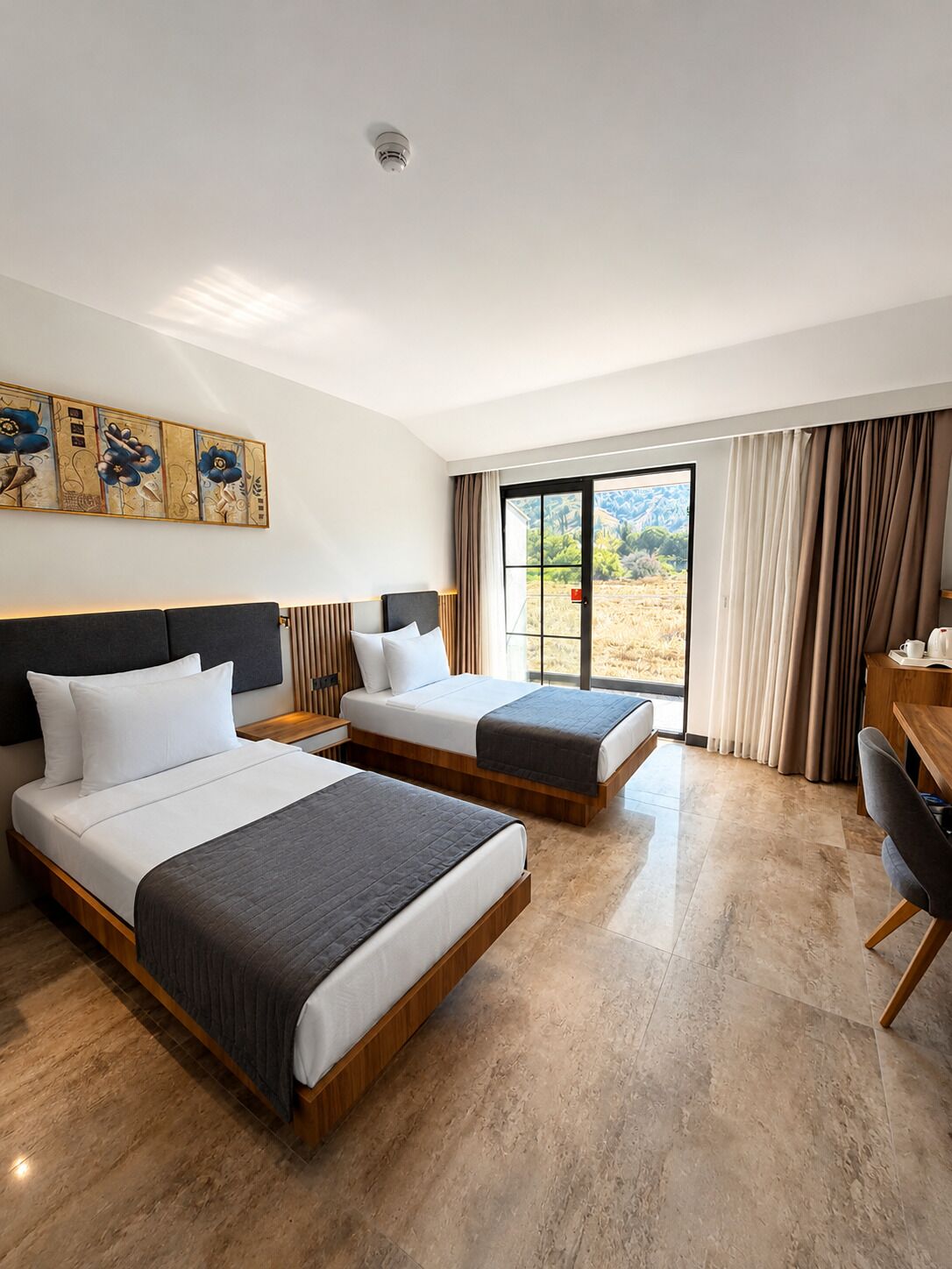 Habitación de hotel con dos camas, un escritorio, una silla, un ventanal con cortinas y una lámpara en el techo.