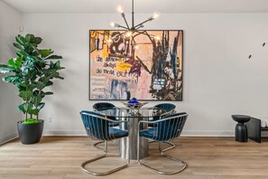A modern dining area with a round table, four chairs, a potted plant, and a large abstract painting on the wall.