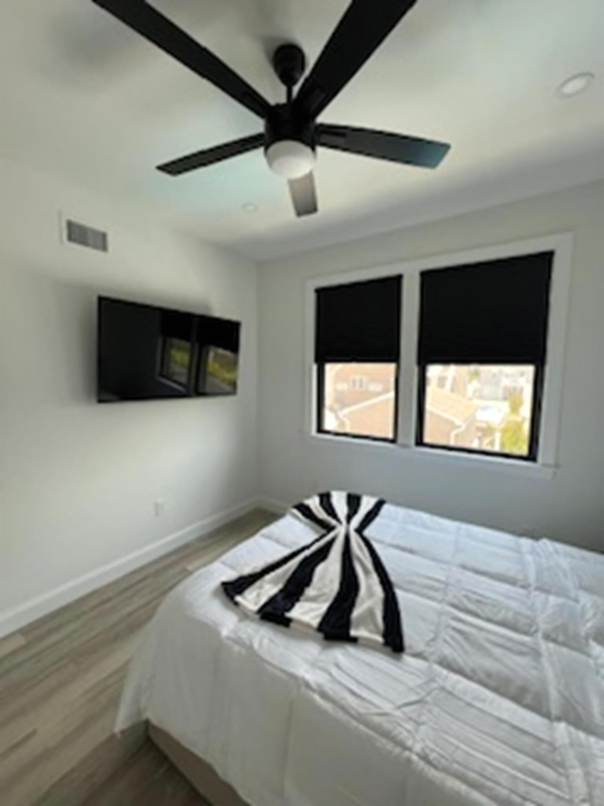 A bedroom with a ceiling fan, a bed with a striped blanket, a mounted TV, and two windows with blackout curtains.