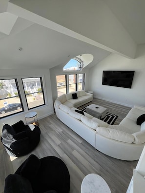 A modern living room with a large L-shaped sectional sofa, a round black chair, a small white side table, and a flat-screen TV mounted on the wall.