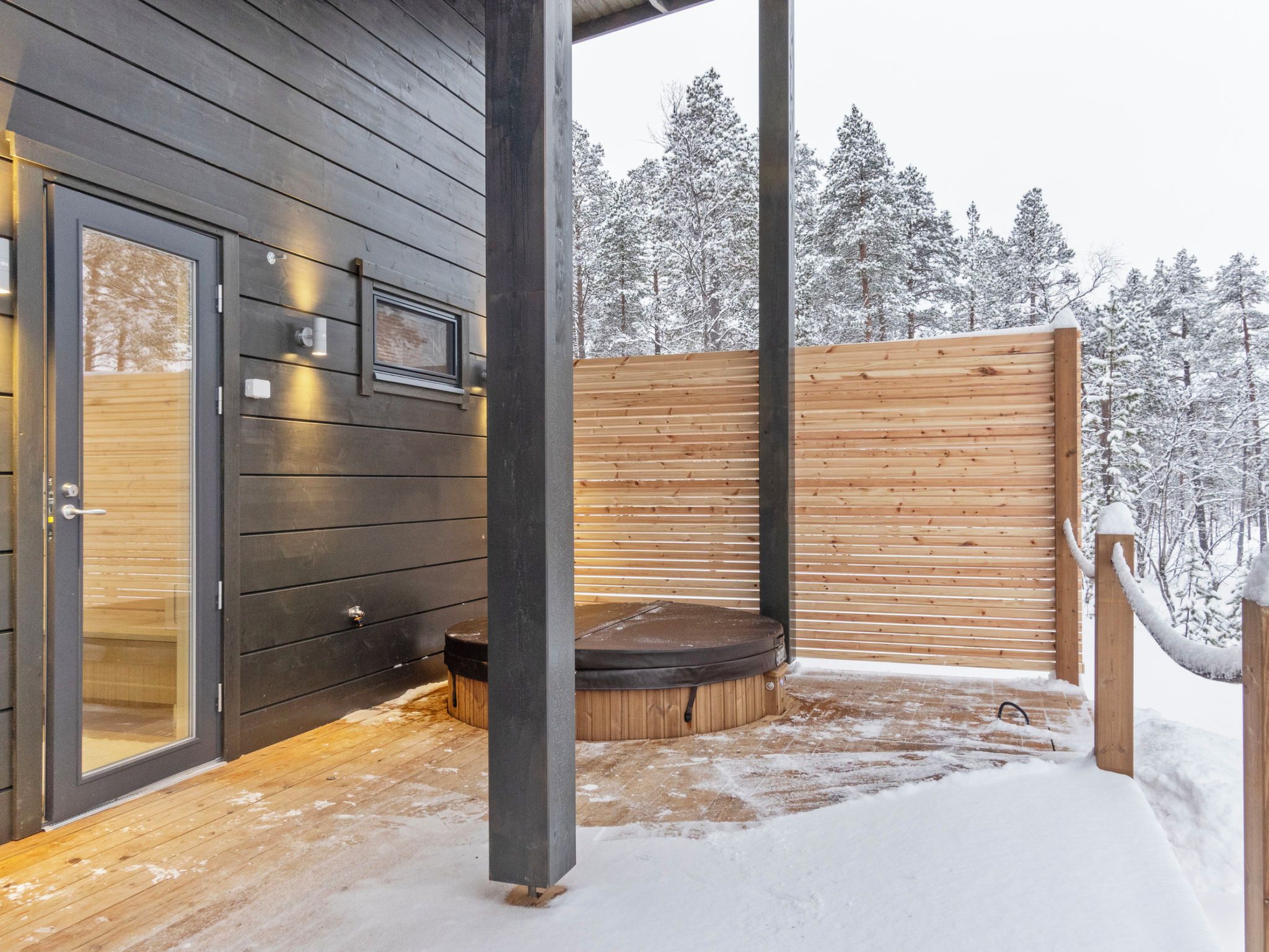 Un área de sauna con un banco de madera y puerta de vidrio, rodeada por una valla de madera y árboles cubiertos de nieve.
