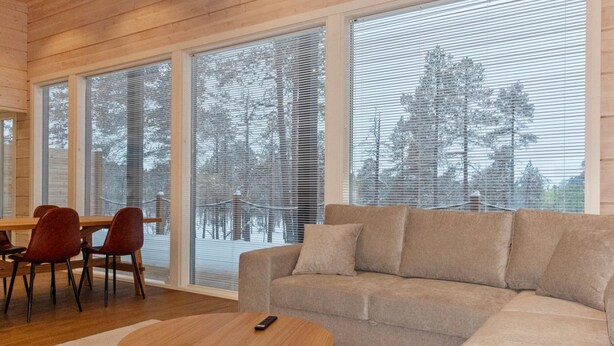 A modern living room with a sofa, a round coffee table, and a dining area with chairs. Large windows offer views of trees and a snowy landscape.