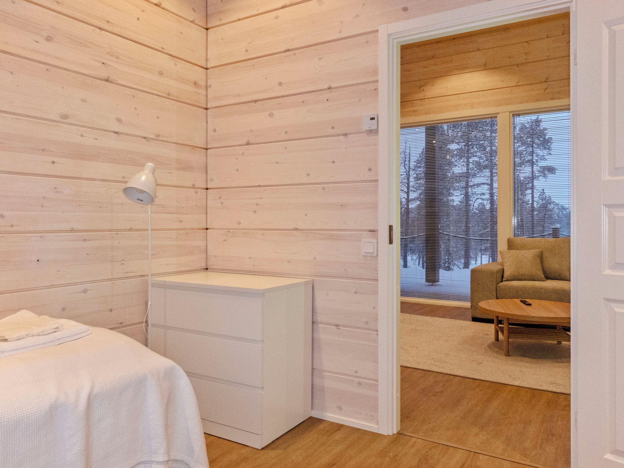 A room with wooden walls, a white bedside table, a lamp, and a view of a sofa and window with blinds.