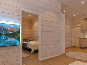 A room with wooden walls, a bed with a white bedspread, a small bedside table, a nightstand, a wall-mounted TV, and a bathroom with a mirror and cabinet. - Villa paadari by Interhome (Inari)