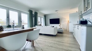 Ein modernes Wohnzimmer mit einer Couch, einem Essbereich mit Tisch und Stühlen, einer Küche mit Schränken und einer Mikrowelle, sowie großen Fenstern mit Vorhängen.