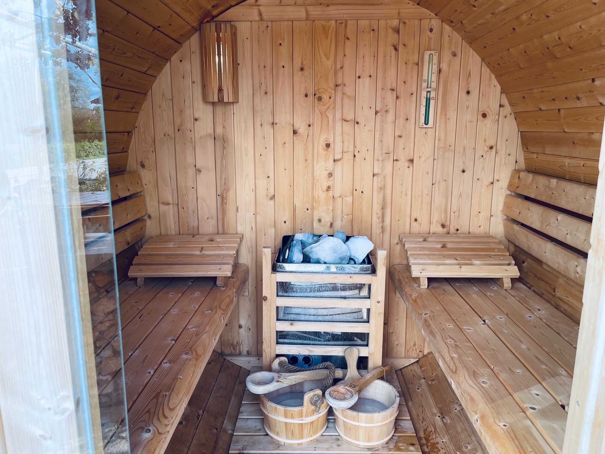 A wooden sauna with benches, a wooden bench with a metal container, and a thermometer on the wall.