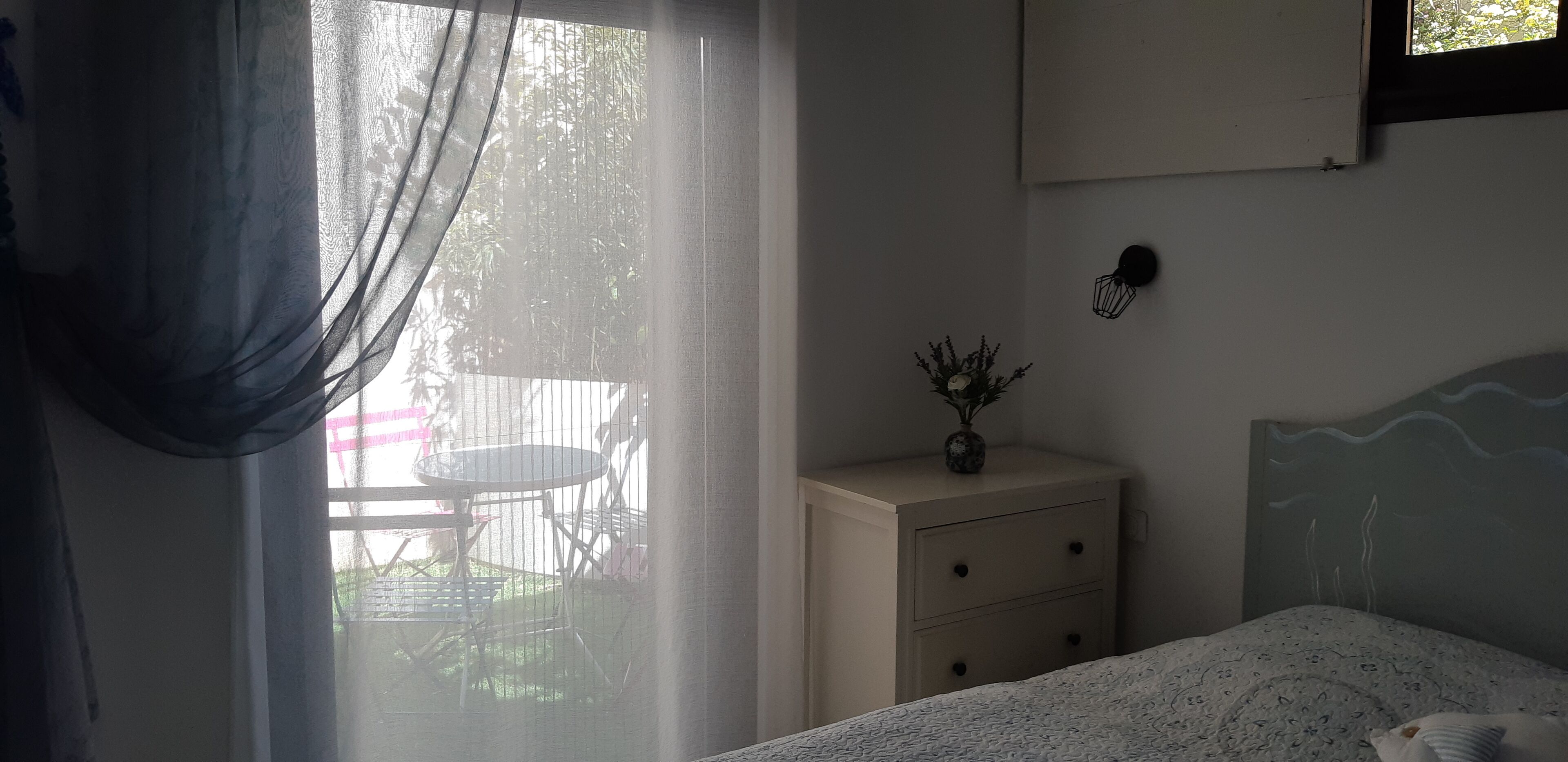 Ein Schlafzimmer mit einem Bett, einem Nachttisch mit einer Vase, einem Fenster mit transparenten Vorhängen und Blick ins Freie.