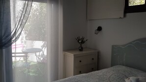 A bedroom with a bed, a bedside table with a vase, a window with sheer curtains, and a view of the outdoors.