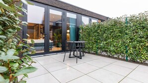 Un bâtiment moderne doté de grandes fenêtres en verre, d’un patio avec une table et des chaises, et d’une haie verdoyante.