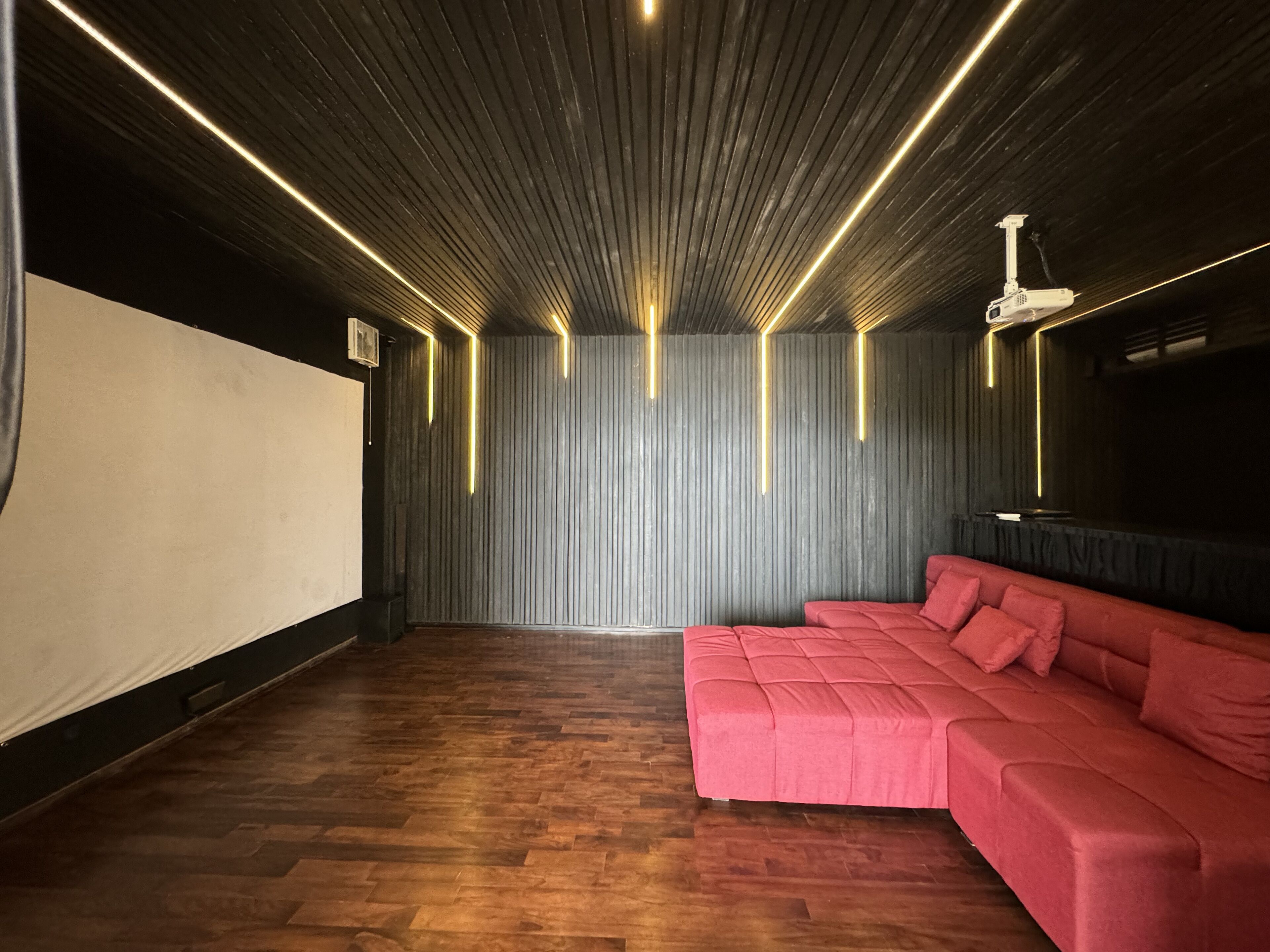 Una stanza con pavimento in legno, un divano rosso, uno schermo per proiettore e un soffitto scuro con luci a LED verticali.