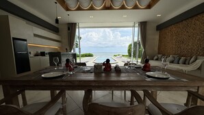 A dining area with a view of the ocean, a kitchen, and a seating area with a stone wall. - Beach front 2 bedroom with cinema room & infinity pool Sahadewa (Bali)