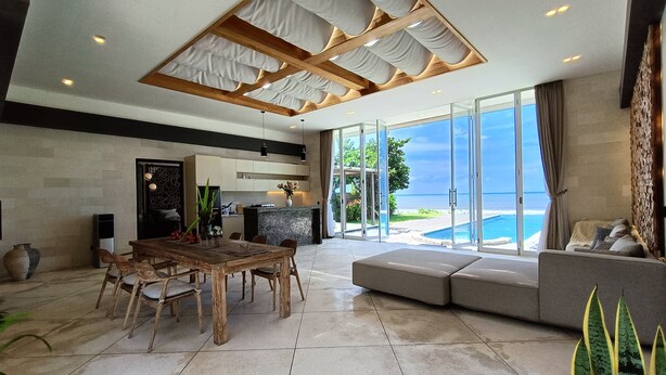 A modern living room with a large glass door leading to a pool, a wooden dining table, and a cozy seating area. - Beach front 2 bedroom with cinema room & infinity pool Sahadewa (Bali)
