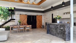 A modern kitchen with a marble island, wooden cabinets, and a dining area with a table and chairs. - Beach front 2 bedroom with cinema room & infinity pool Sahadewa (Bali)