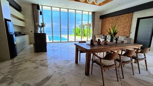 A spacious dining area with a large table and chairs, a kitchen with a refrigerator and cabinets, and a view of the ocean through large sliding doors. - Beach front 2 bedroom with cinema room & infinity pool Sahadewa (Bali)