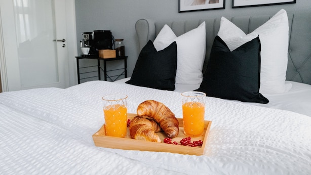 Ein ordentlich bezogenes Bett mit weißer Bettwäsche, ein Tablett mit Croissants und Orangensaft sowie eine Kaffeemaschine auf einem Beistelltisch.