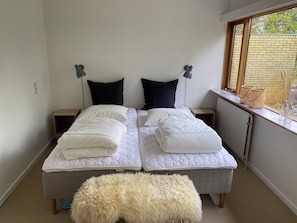 Een tweepersoonsbed met witte beddengoed, twee nachtkastjes met lampen, een raam met uitzicht op groen, en een zachte witte poef.