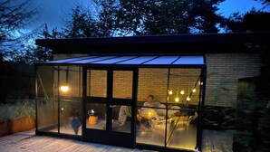 A modern glass-enclosed structure with a wooden deck, brick walls, and a visible interior with seating and lighting.