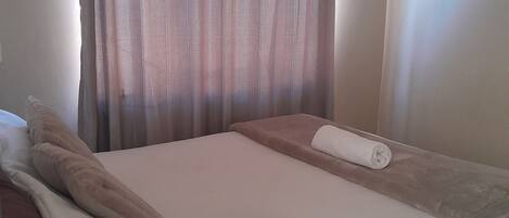 A neatly made bed with a white duvet, a brown throw blanket, and a rolled-up towel.