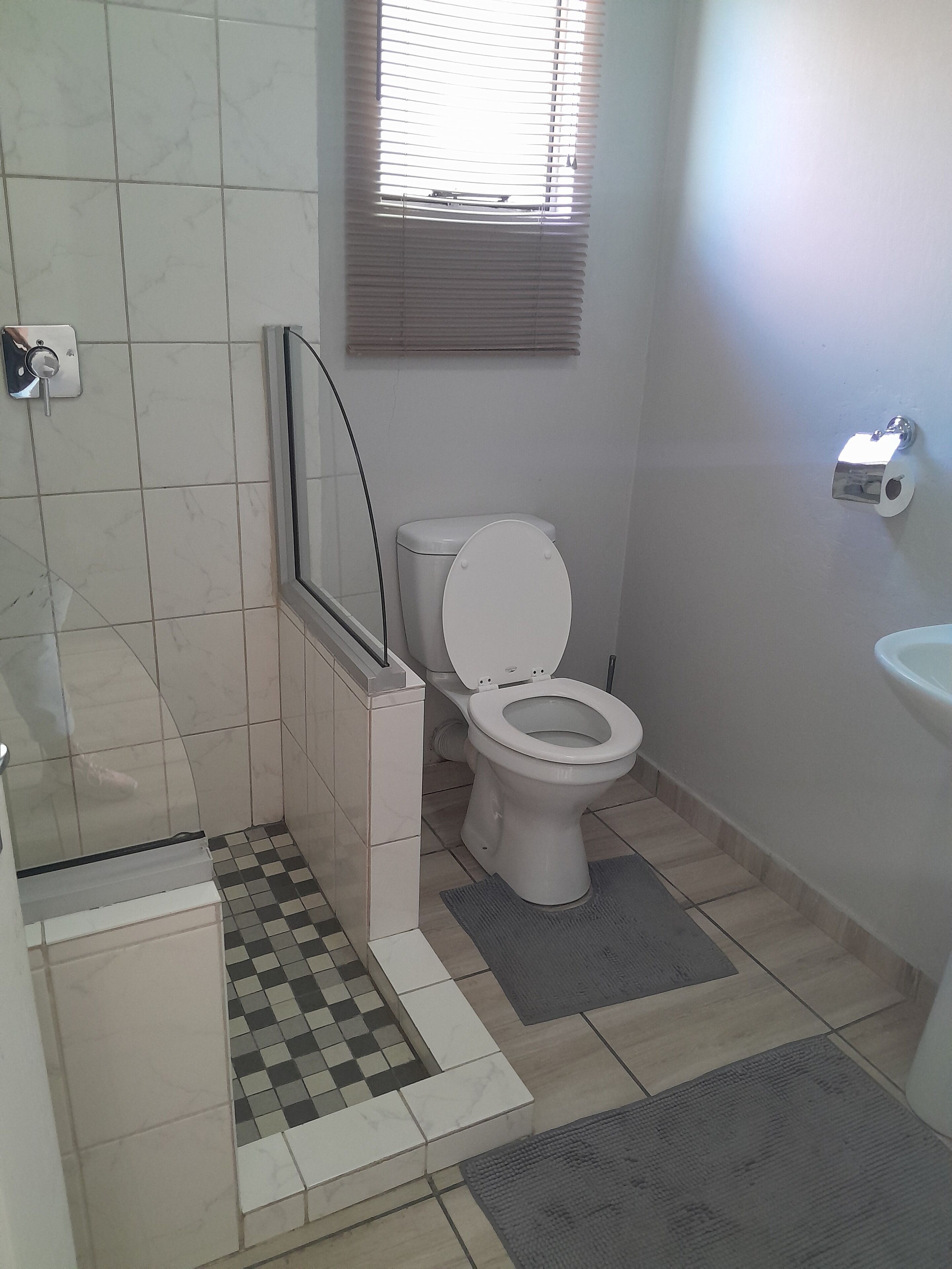 Un baño con inodoro, lavamanos, zona de ducha y ventana con persianas.