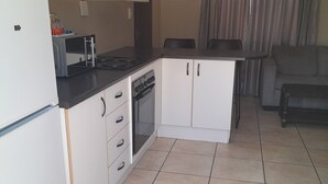 A compact kitchen with white cabinets, a built-in oven, and a microwave. There is a dining area with a table and chairs, and a grey sofa in the living room. - High Breeze Lodge (Upington)