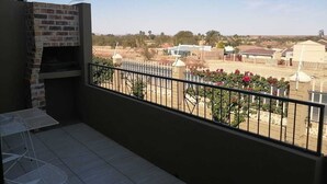 A balcony with a brick fireplace, a metal table, and a view of a residential area with houses and greenery. - High Breeze Lodge (Upington)
