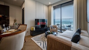 Un salon moderne doté d’un grand téléviseur à écran plat affichant le logo « NETFLIX », d’un coin repas avec une table en bois et d’un canapé offrant une vue sur la ville grâce à de grandes fenêtres.