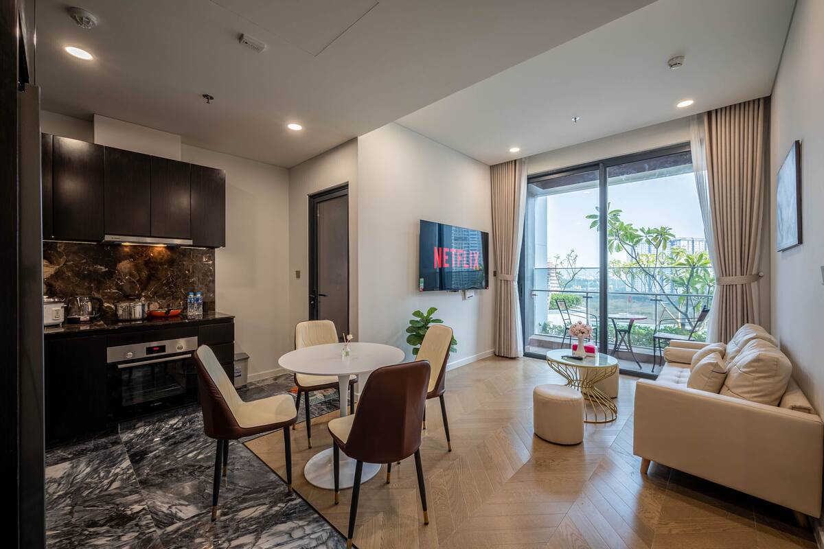 A modern living room with a dining area, a kitchen with dark cabinets, a flat-screen TV, and a large window with a view.