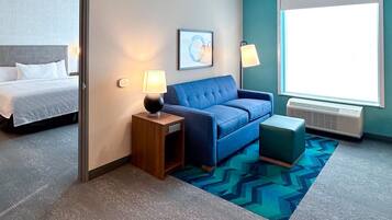 A hotel room with a bed, a blue sofa, a small table with a lamp, a picture on the wall, and a large window.