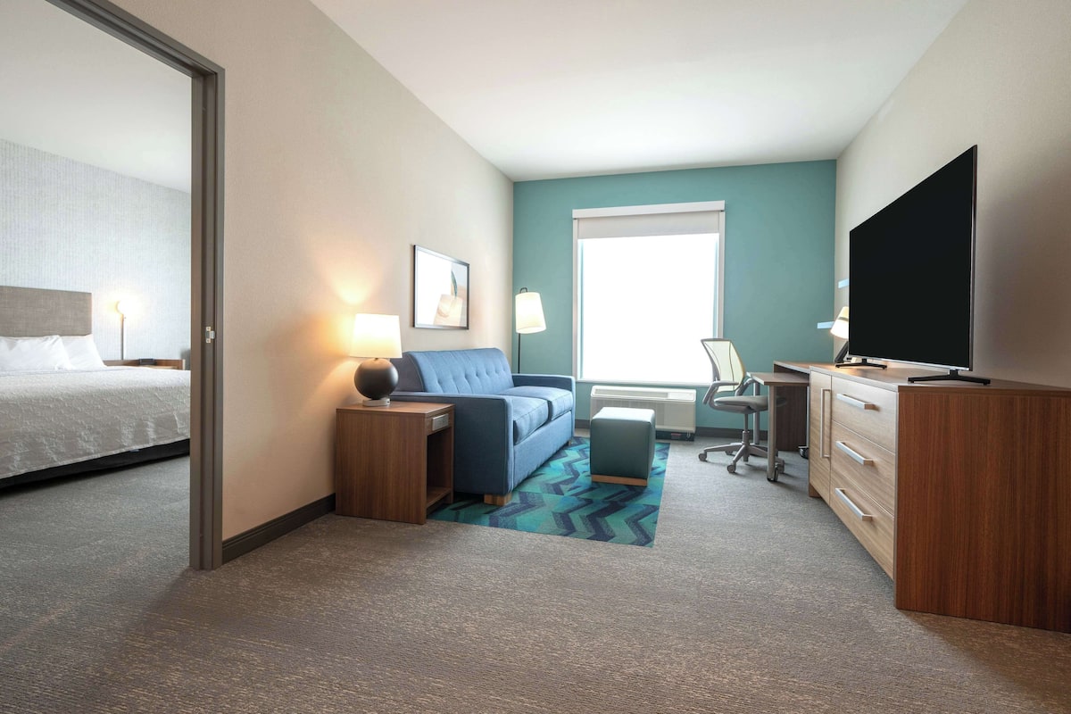 Habitación de hotel con cama, sofá azul, escritorio con computadora, televisor de pantalla plana y ventana con persianas.