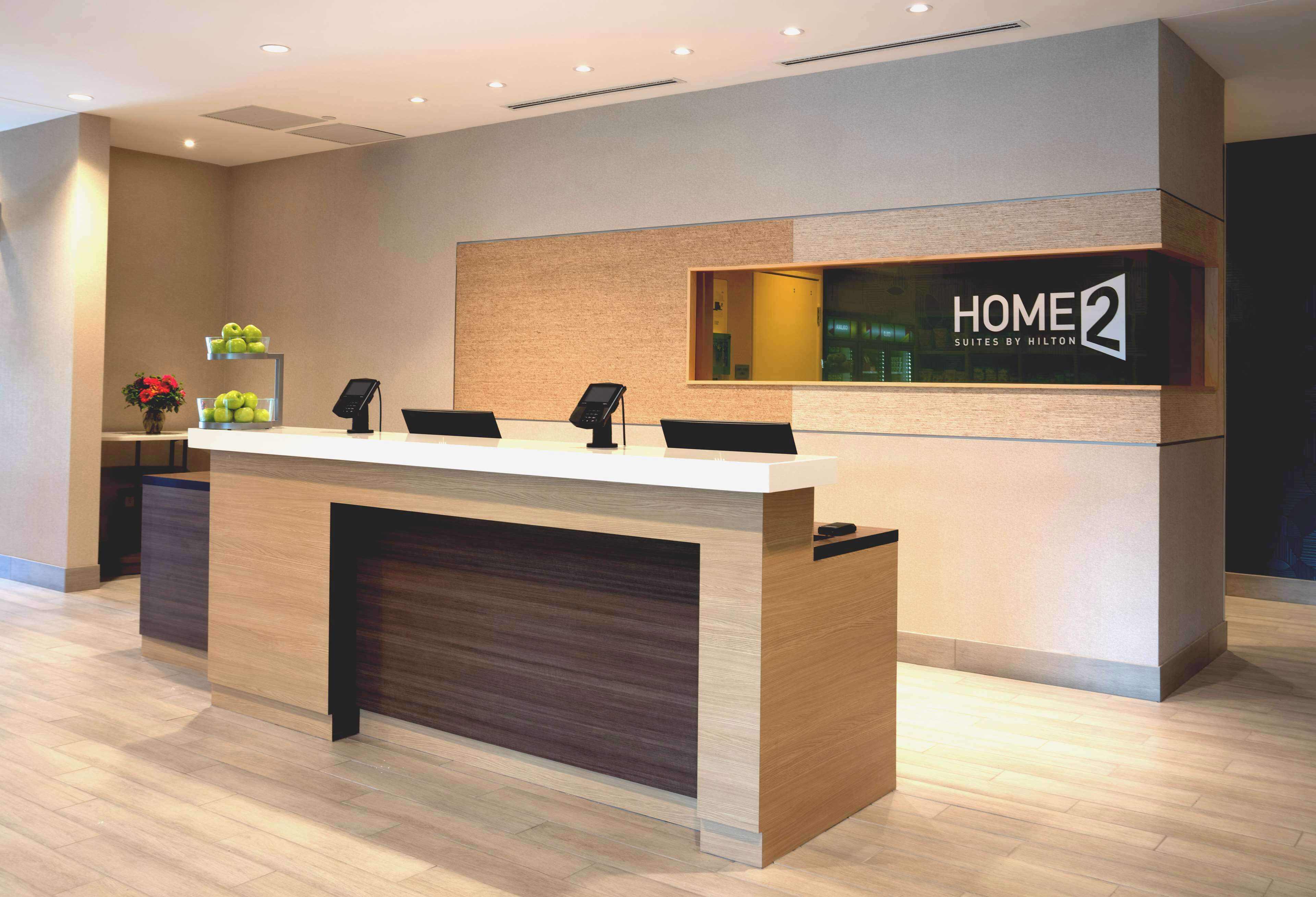 Hotellin vastaanottotiski, jossa on kyltti, jossa lukee ”HOME 2 Suites by Hilton”, puinen tiski ja kukkakimppu.