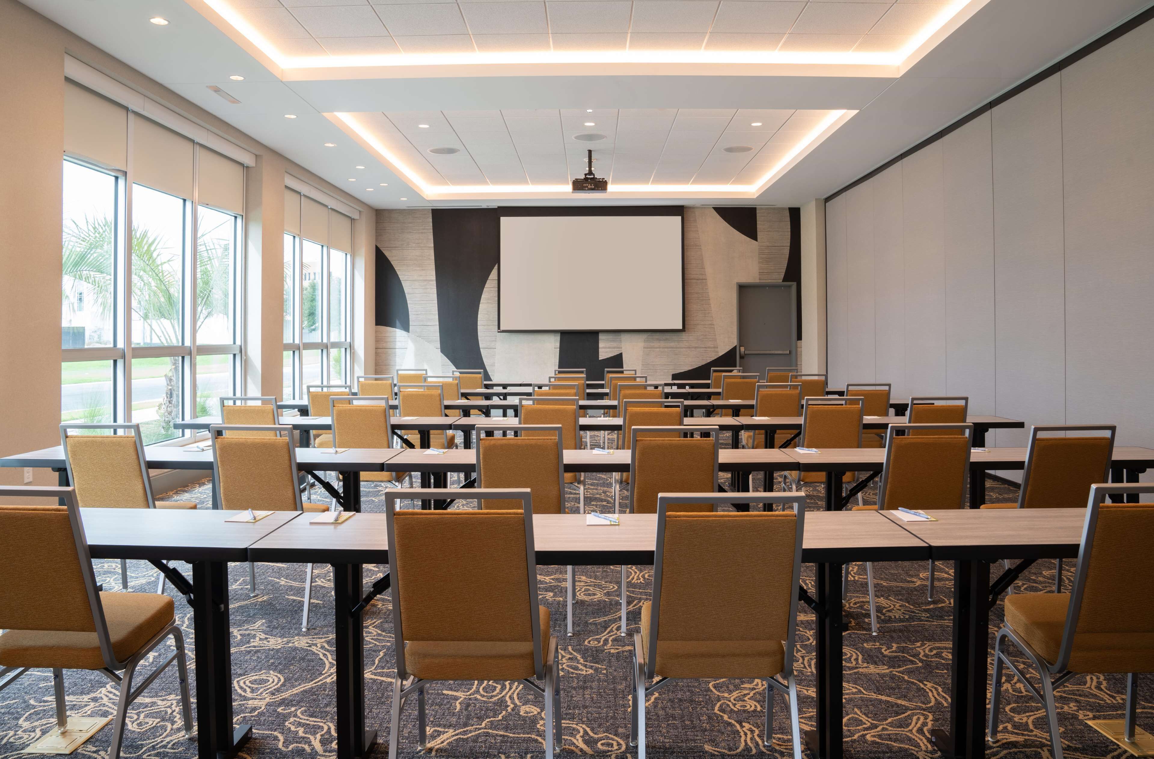 A conference room with multiple tables and chairs, large windows, and a projector screen.