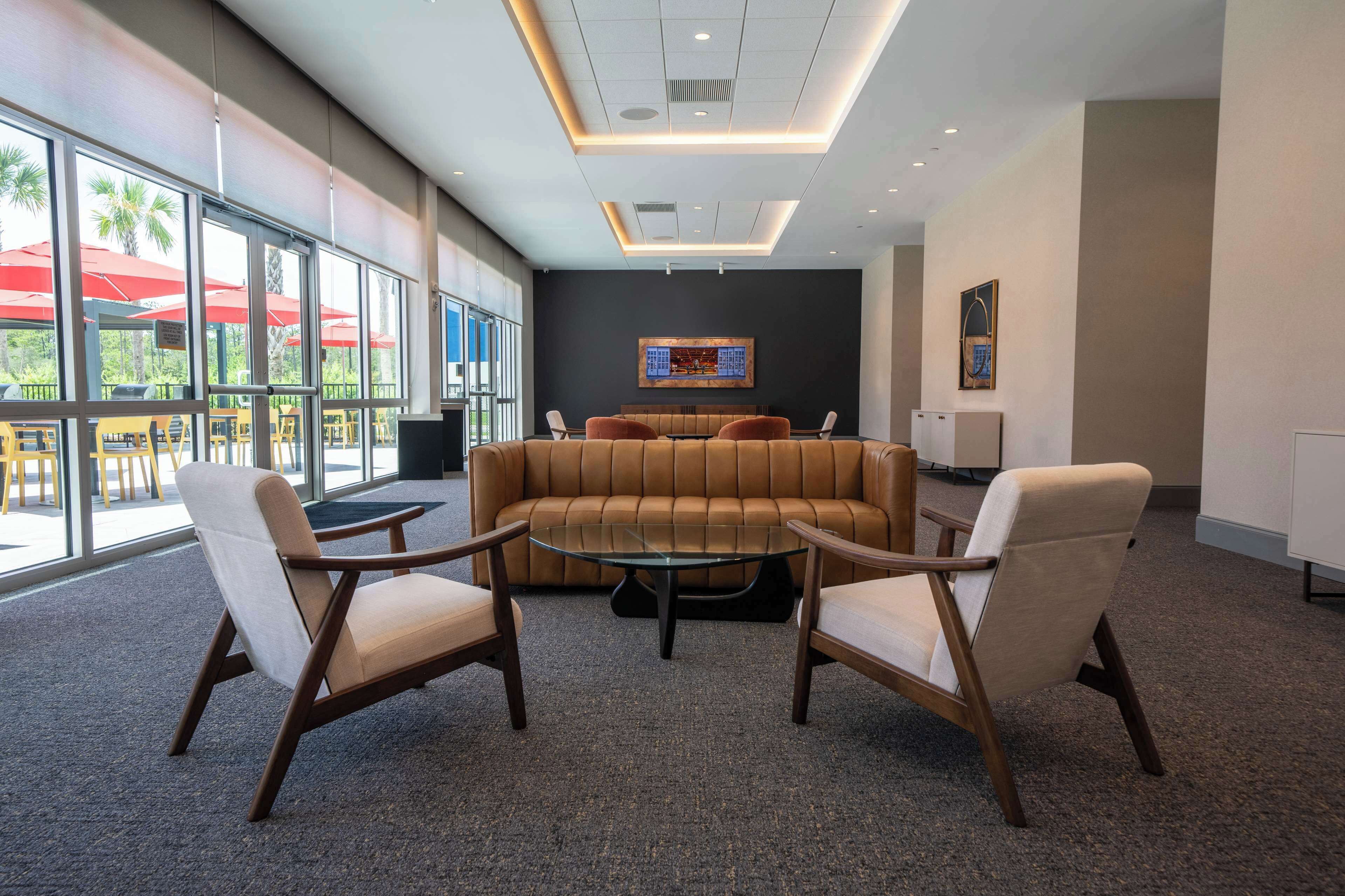 Un área de descanso moderna con un sofá de cuero, dos sillones y una mesa de centro de vidrio. Amplias ventanas ofrecen vistas a un espacio exterior con asientos.