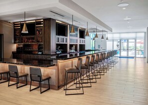 A modern bar area with a long counter, multiple bar stools, and pendant lighting. - Home2 Suites by Hilton Fort Walton Beach Eglin AirForce Base (Fort Walton Beach)
