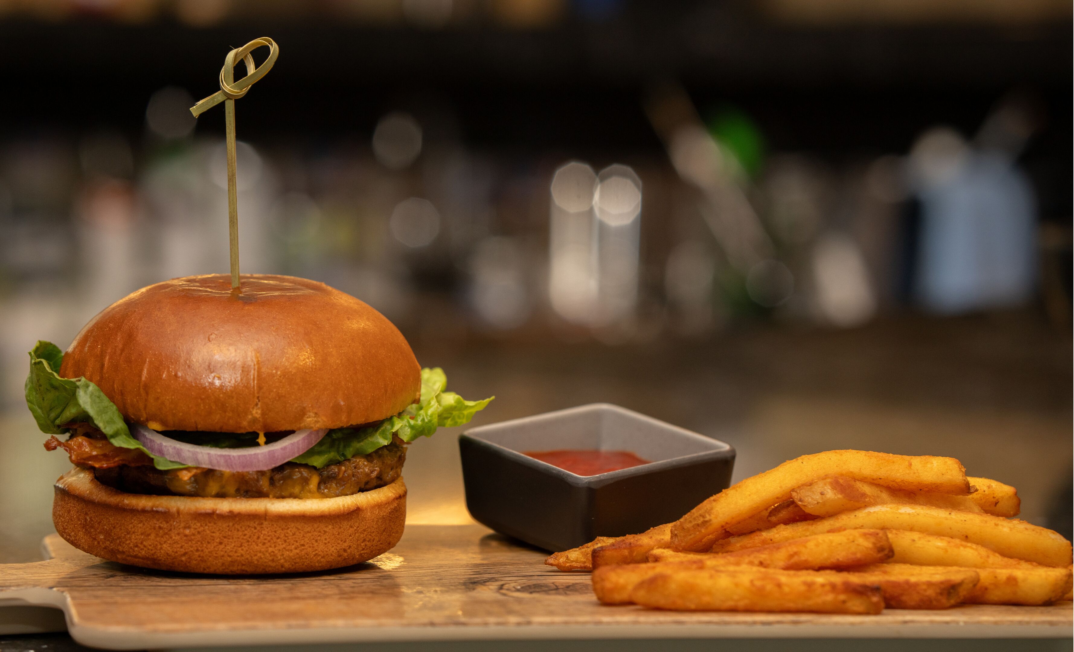 A burger with a toothpick, lettuce, onion, and a bun, alongside a small bowl of ketchup and a serving of fries.