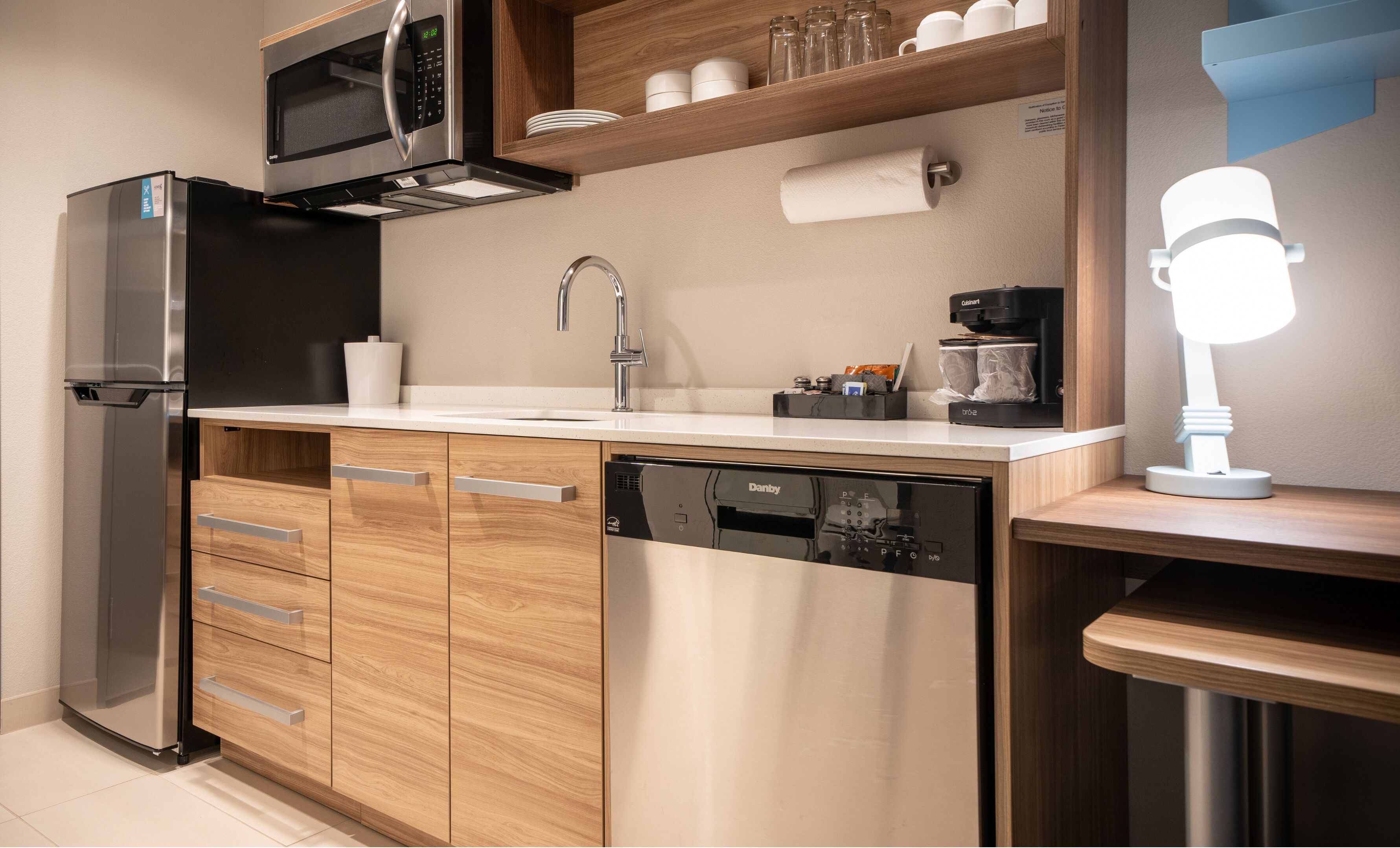 A modern kitchen with a stainless steel refrigerator, a dishwasher, wooden cabinets, a microwave, a coffee machine, and a bright light fixture.