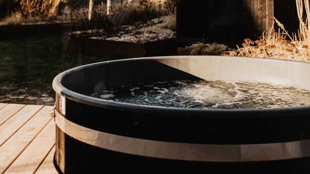 Un jacuzzi en una terraza de madera con vistas a un edificio moderno y un árbol sin hojas al fondo.