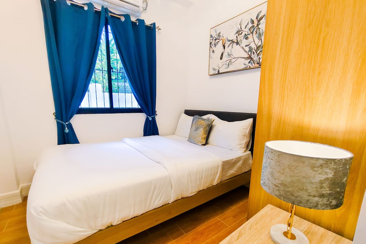 A single bed with a wooden frame, a bedside table with a lamp, and a window with blue curtains.