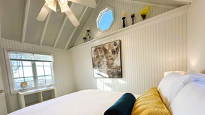 Een slaapkamer met een groot bed, een raam met zonwering, een plafondventilator en een kunstwerk aan de muur.