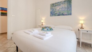 Un lit bien fait, avec du linge de lit et des serviettes blanches, un tableau représentant un paysage, et deux tables de chevet avec des lampes.