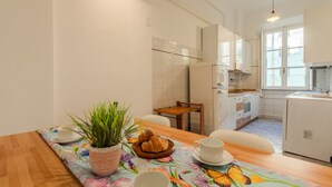 Une cuisine moderne avec des meubles blancs, une table à manger recouverte d’une nappe à motifs floraux et une plante en pot.