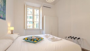 Un letto rifatto con cura, un cuscino colorato, una lampada da comodino, una finestra con vista sugli edifici e uno stendibiancheria.
