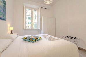 Un lit bien fait, agrémenté d’un coussin coloré, une lampe de chevet, une fenêtre donnant sur des immeubles et un étendoir.