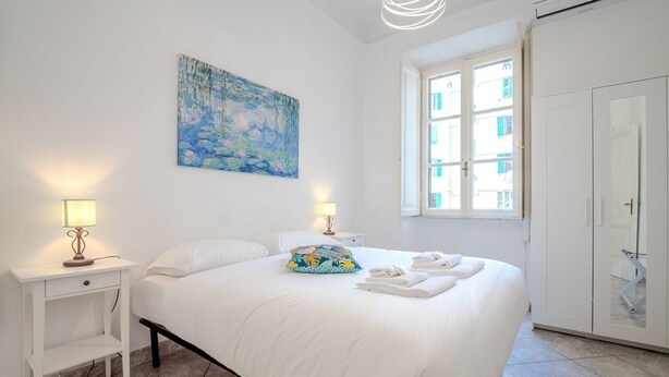 Un lit bien fait, avec des draps blancs, un coussin coloré et des serviettes pliées. Il y a un tableau au mur, une fenêtre avec des stores et une armoire.
