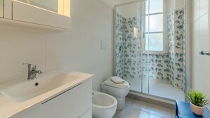 Une salle de bain moderne avec un lavabo blanc, deux toilettes, une douche en verre avec des rideaux à motifs floraux et une petite plante en pot.