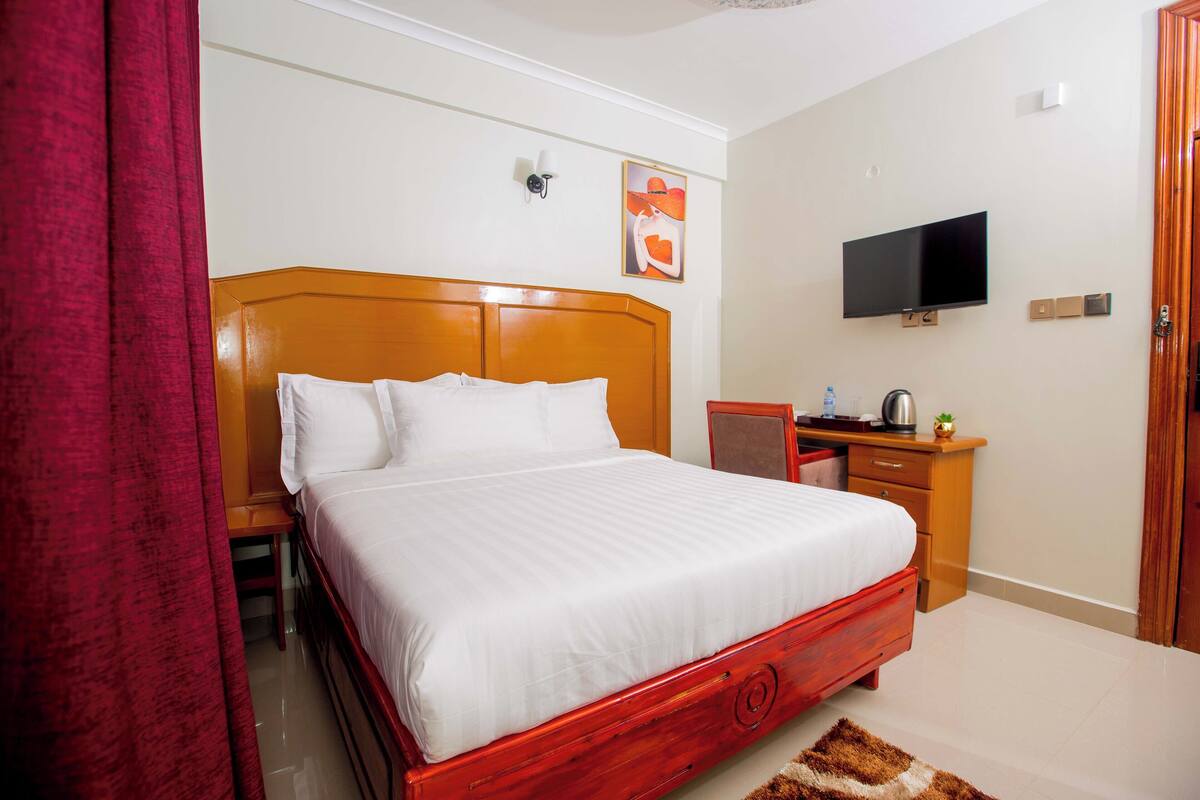 A hotel room with a wooden bed, a television mounted on the wall, a desk with a chair, and a red curtain.