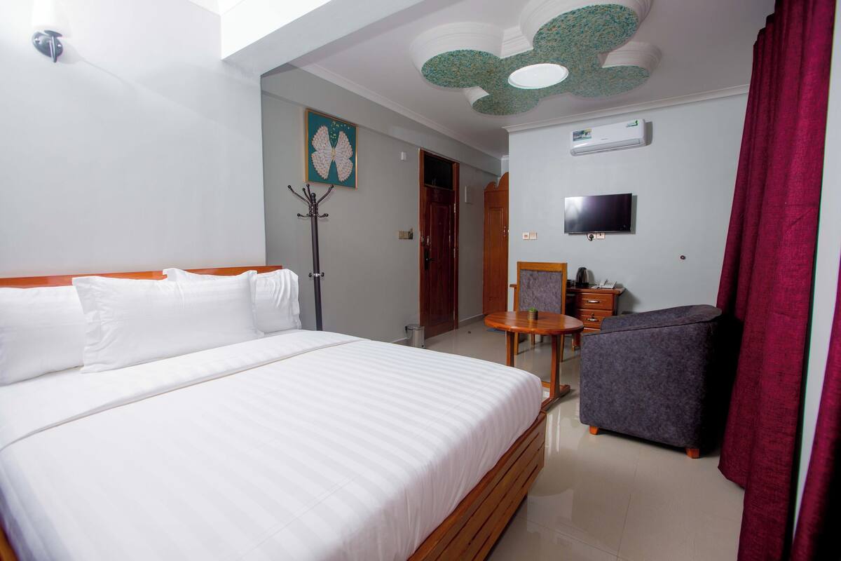 A hotel room with a large bed, a small table, a chair, a TV, and a ceiling fan.
