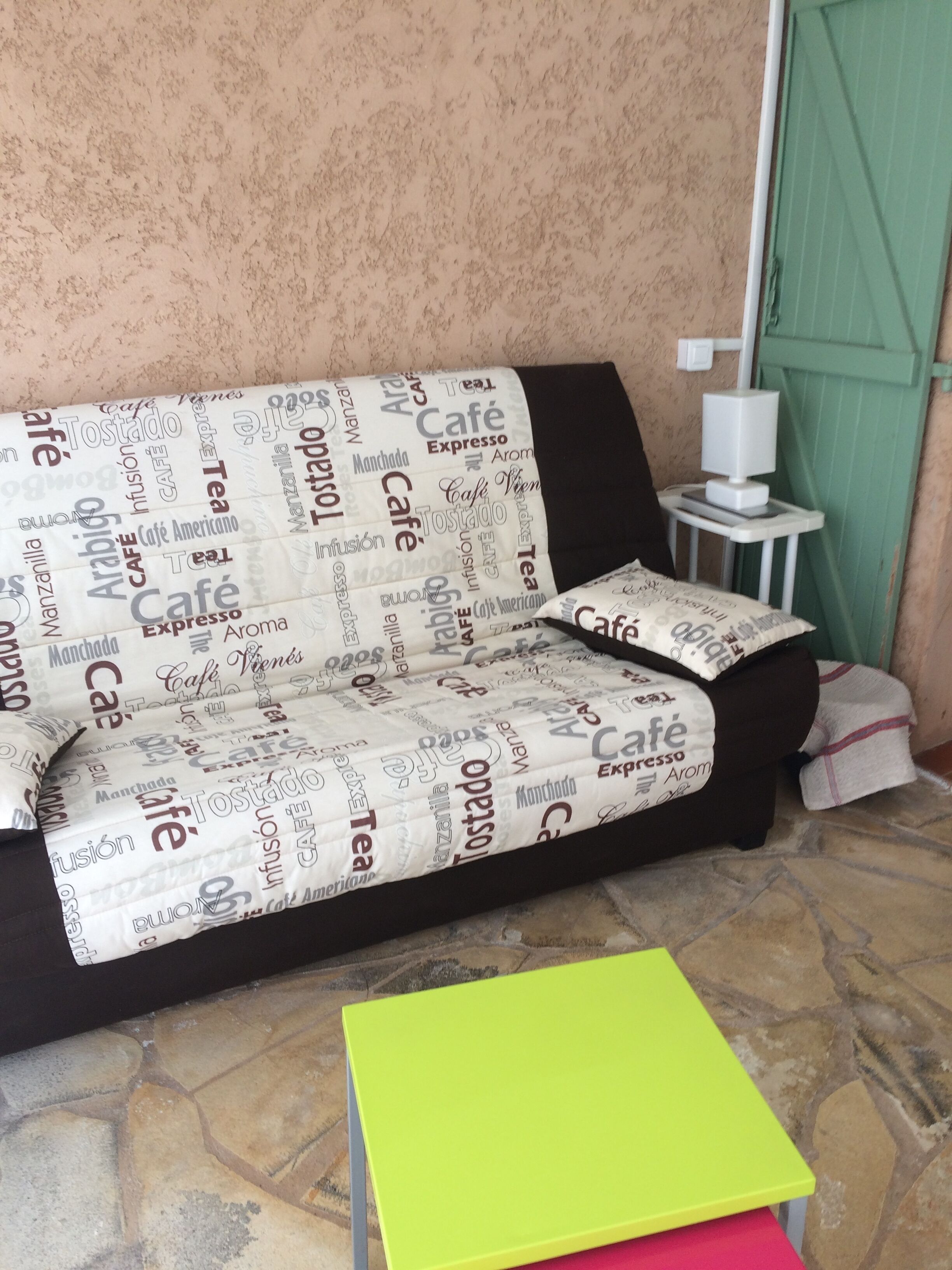 Un canapé recouvert d’un tissu à motifs, une petite table blanche et une boîte verte posée sur un sol en pierre.